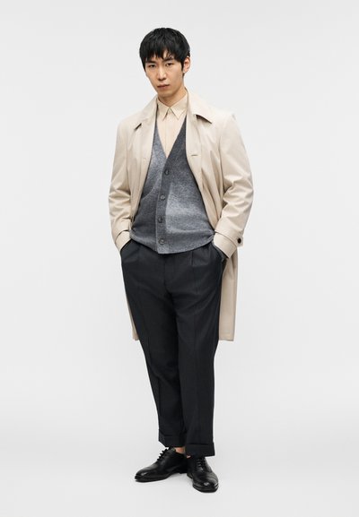 Μπεζ καμπαρντίνα πάνω από γκρι ζακέτα, κρεμ πουκάμισο και σκούρο παντελόνι με λεπτές ρίγες. Μαύρα παπούτσια ολοκληρώνουν την εμφάνιση. Απλός, ραμμένος σχεδιασμός.