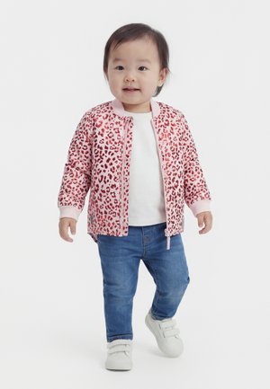 Lille barn iført en pink leopardmønstret jakke, hvid skjorte, blå jeans og hvide sneakers, stående og smilende på en hvid baggrund.