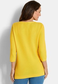 Gelber Strickpullover mit Dreiviertelärmeln, weitem Ausschnitt und geradem Saum. Weiche Textur mit gerippter Verarbeitung an den Bündchen und am Saum.