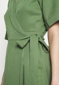Abito avvolgente verde con maniche corte, dotato di una cintura in vita, tessuto strutturato e silhouette aderente. Nessun motivo visibile.