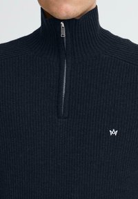 Maglione scuro a maglia con colletto alto e mezza zip, caratterizzato da una trama a costine e un piccolo logo bianco.