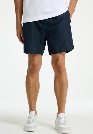 Mees kannab tumesiniseid lühikeseid pükse, valgeid tossusid ja valget särki, käed taskutes, seisab valgel taustal.