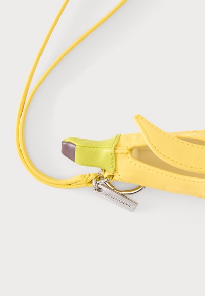 Sac bandoulière en cuir jaune en forme de banane avec une pointe verte et marron, anneau porte-clés argenté et longue bandoulière sur fond blanc.