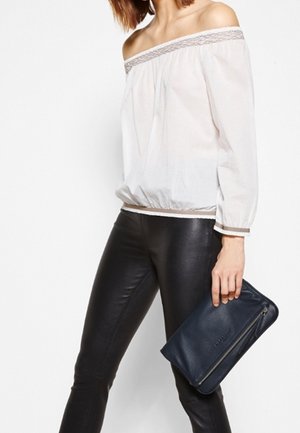 Femme portant un blouse blanche à épaule dénudée et un pantalon en cuir noir, tenant une pochette zippée bleu marine dans sa main droite.