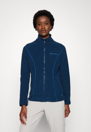 Frau trägt eine marineblaue Jacke mit Reißverschluss, hohem Kragen und Seitentaschen, kombiniert mit hellen Hosen, stehend vor einfachem Hintergrund.