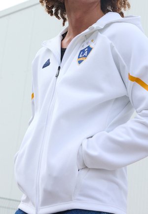 Osoba w białej kurtce na zamek LA Galaxy z logo Adidas i złotym paskiem na rękawie, z rękami w kieszeniach, na zewnątrz.