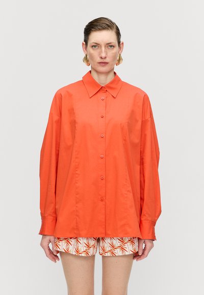 Bright orange button-up shirt made from a smooth fabric, featuring a pointed collar and long sleeves, paired with patterned white shorts.