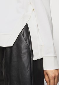 Bílá mikina s žebrovanou texturou a bočními rozparky, doplněná hladkými černými koženými kalhotami. Viditelné švy a volný střih.