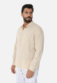 Camicia a maniche lunghe in lino beige con colletto a band, con chiusura a bottoni e dettagli ai polsini, abbinata a pantaloni bianchi.