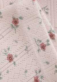 Stickad textil i mjuk rosa med små blommönster i maroon och grönt, med en texturerad design och öppna vävda sektioner.