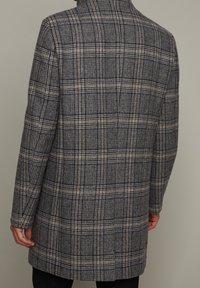 Cappotto scozzese in blu scuro, beige e grigio. Forma su misura con colletto strutturato e maniche lunghe. Il tessuto appare testurizzato e caldo.