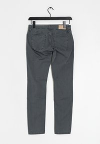 Jeans ajustados grises con una textura suave, con bolsillos traseros y un sutil detalle de costura, colgados de un gancho negro sobre un fondo blanco.