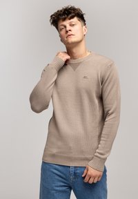 Beżowy sweter dzianinowy z ryflowanym dekoltem i mankietami, z małym logo na piersi. Model pozujących w niebieskich dżinsach.