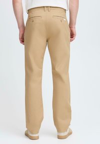 Beige cotton trousers with straight legs, featuring back pockets and a tag; worn with light-colored sandals on a plain background.