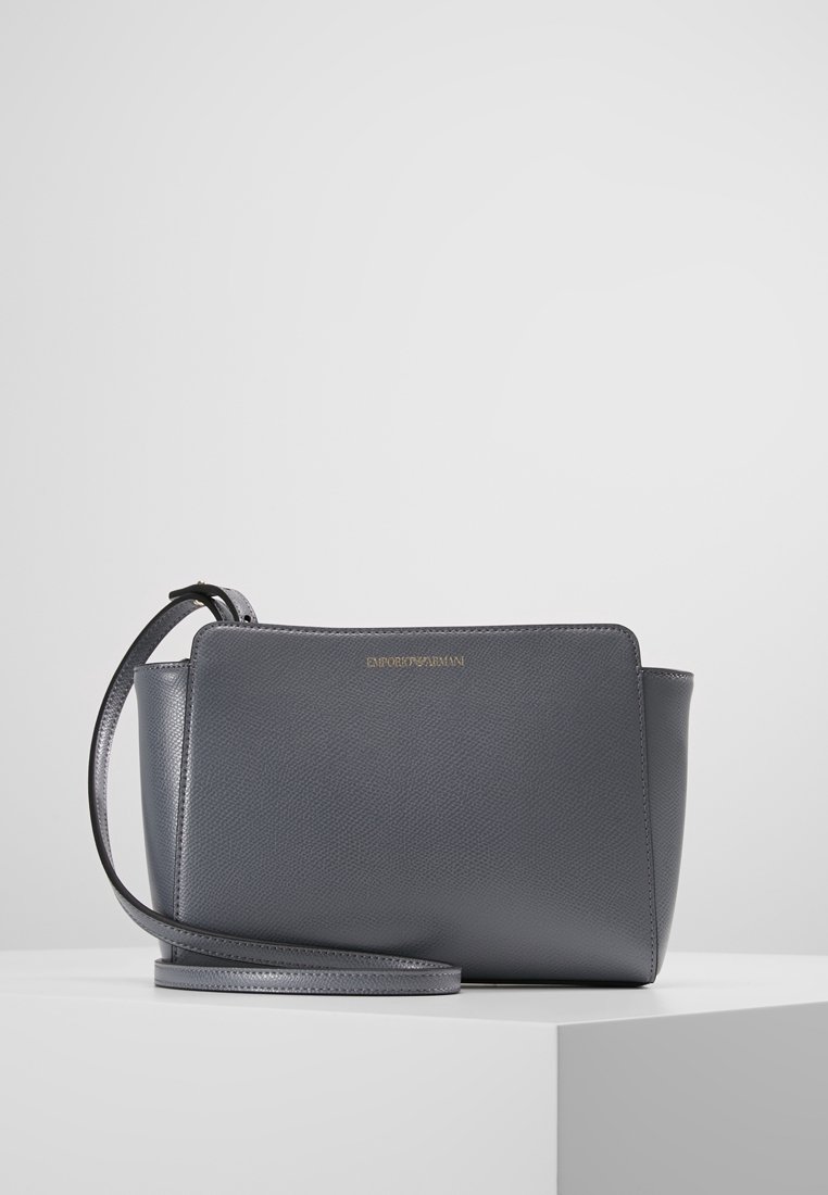 Szara skórzana torebka o strukturalnym kształcie, z płaskim dnem, regulowanym paskiem i wytłoczonym logo z przodu. Gładka tekstura.