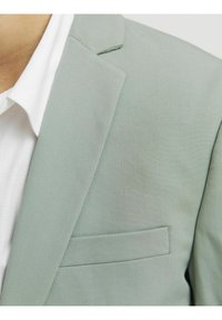Jasnozielona marynarka z teksturowanej tkaniny, z wykładanym kołnierzem i kieszenią na piersi. Nosi się ją na białej koszuli z kołnierzykiem.