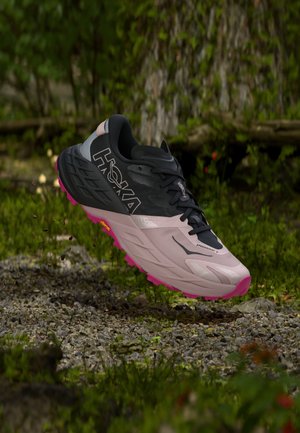 Trailrunning-Schuh mit pinker Sohle und hellrosa-schwarzem Obermaterial schwebt über einem Kiesweg im Wald.