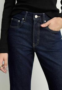 Jean bleu foncé avec des coutures jaunes contrastantes, poches avant et fermeture à bouton. Le tissu a une texture lisse en denim.