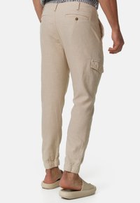 Pantalones cargo de lino beige con puños elásticos y bolsillos traseros, que presentan un corte relajado y detalles de diseño utilitarios.