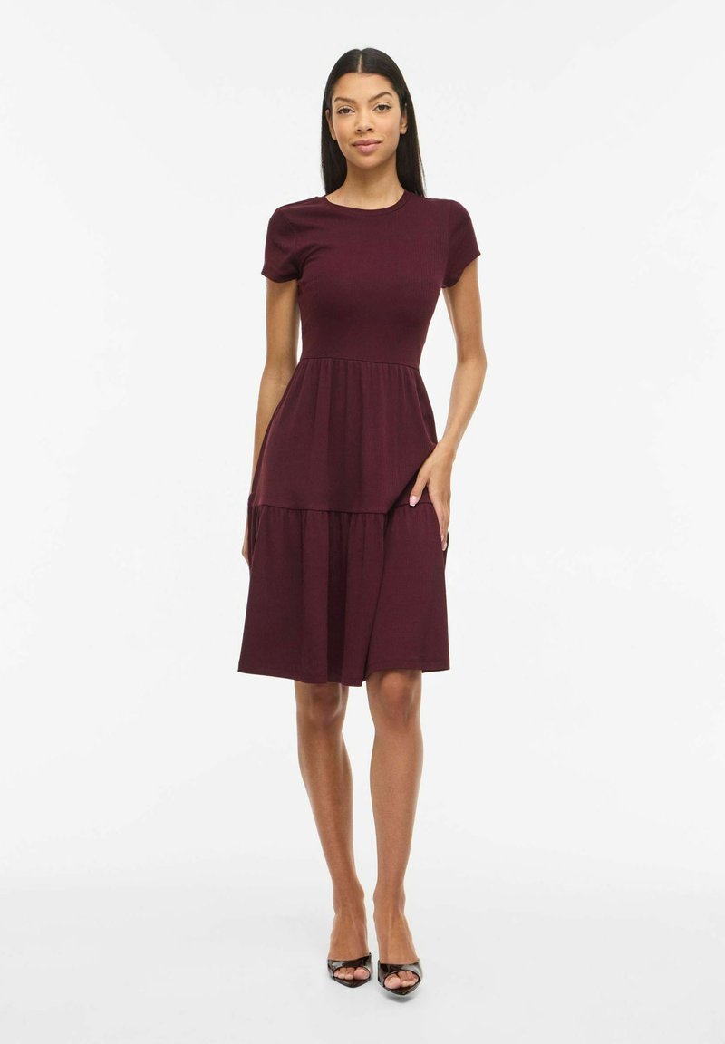 Žena stoji okrenuta prema naprijed, nosi bordo haljinu s kratkim rukavima do koljena i crne cipele s otvorenim prstima na bijeloj pozadini.