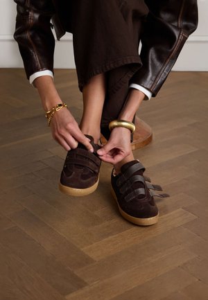 Bruine suède sneakers met drie klittenbandsluitingen en een getextureerde tan rubberen zool, gedragen door een persoon die ze vastmaakt terwijl hij op een houten vloer zit.