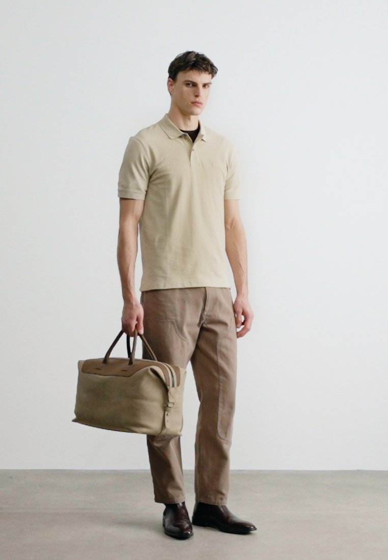 Un homme portant un polo beige et un pantalon marron, tenant un grand sac de voyage marron par les poignées, debout contre un mur blanc uni.