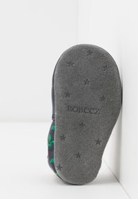 Chaussure à semelle souple grise avec une surface texturée, présentant des motifs de dinosaures verts sur une tige navy, marquée "ROBEEZ" sur la semelle.