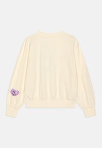 Felpa color crema con maniche a palloncino, polsini a coste e vita aderente. Presenta una toppa a forma di cuore viola con paillettes sulla manica sinistra.