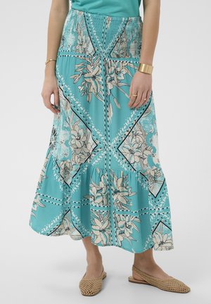 Femme portant une jupe midi turquoise à fleurs avec taille smockée, un haut bleu canard, un bracelet en or, une bague et des ballerines tressées beige sur fond blanc.