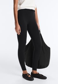 Personne portant un pantalon noir ajusté avec une petite ceinture, des chaussures noires à enfiler, et tenant un grand sac fourre-tout noir.