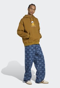 Brun hætte sweatshirts med et grafisk logo på brystet, parret med blå mønstrede brede bukser med et gentaget logo-design og sneakers.