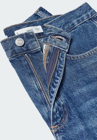 Jean bleu en denim plié avec un bouton métallique visible et une fermeture éclair partiellement ouverte sur un fond clair.