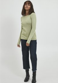 Un haut vert clair à manches longues en tissu doux, associé à un jean cropped bleu foncé avec des ourlets effilochés et des bottines noires.