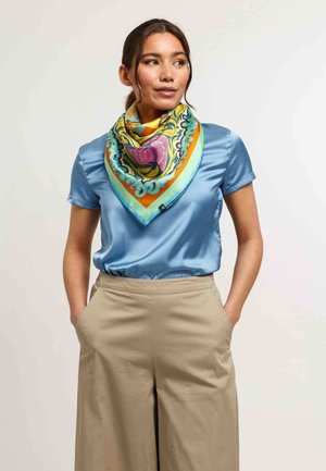 Foulard quadrato in seta multicolore con motivi floreali; indossato su una blusa di raso blu chiaro; presenta vivaci accenti di arancio e verde.