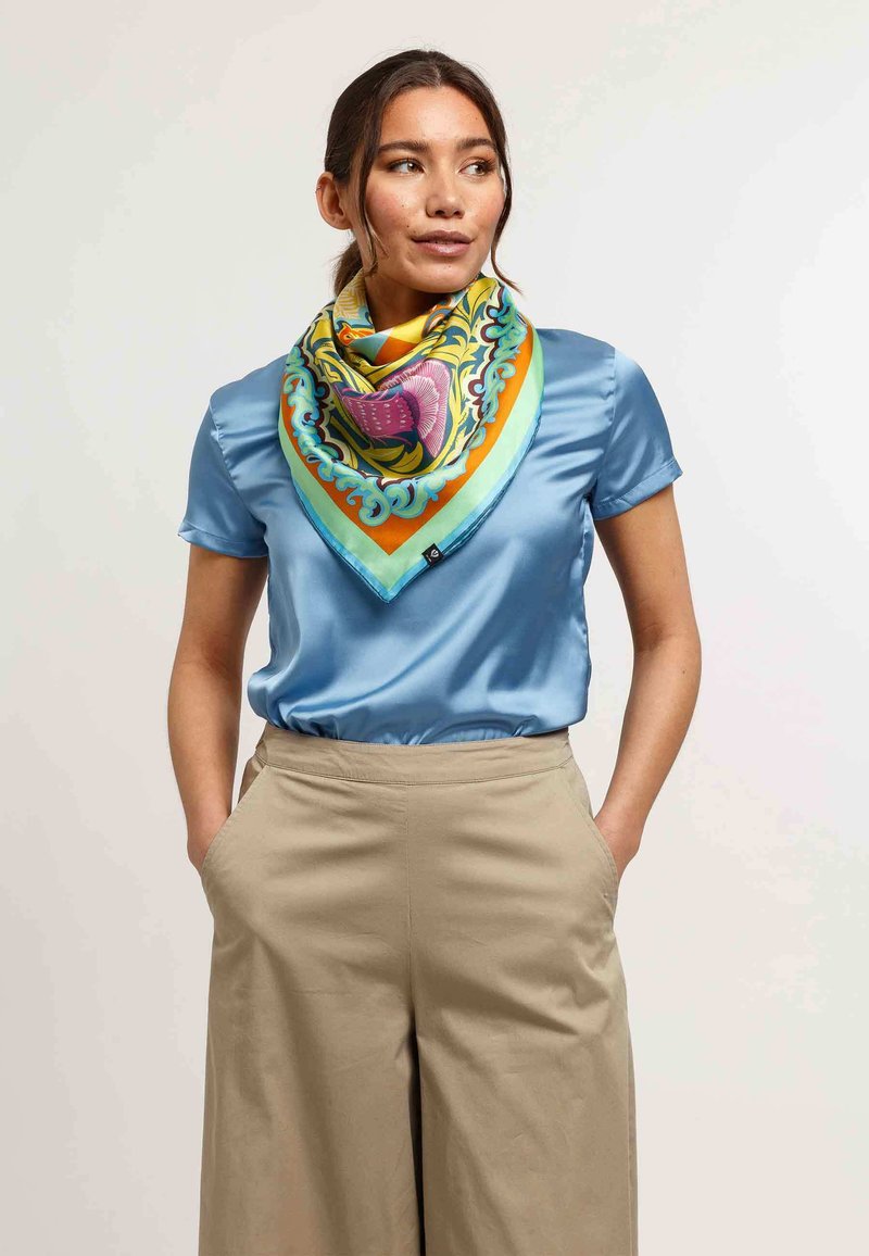 Vierkante, multikleurige zijden sjaal met bloemenpatronen; gedragen over een lichtblauw satijnen blouse; heeft levendige accenten van oranje en groen.