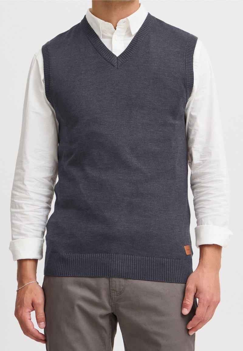 Veste grise en maille avec un col en V, des bords côtelés et une texture plate. Portée sur une chemise blanche à col. Patch en cuir près de l'ourlet.