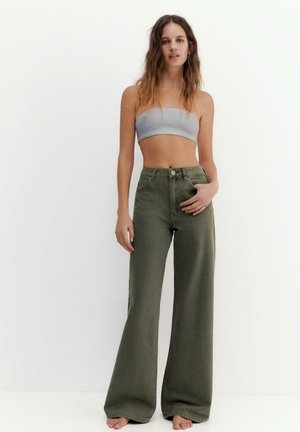 Jeune femme debout, pieds nus, portant un haut bandeau gris et un pantalon taille haute à jambes larges de couleur vert olive, sur un fond blanc uni.