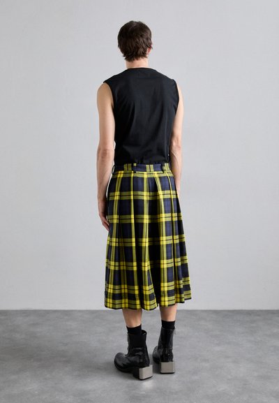Schwarzes ärmelloses Oberteil, kombiniert mit einem knielangen Kilt mit einem gelb-blauen Karomuster; getragen mit schwarzen glänzenden Knöchelstiefeln.