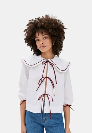 Donna con capelli ricci che indossa una camicetta bianca con un grande colletto e tre lacci frontali abbinata a jeans blu su sfondo neutro.
