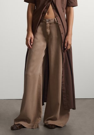 Person trägt beige Hosen mit weitem Bein, Leopardenmuster-Schuhe und ein langes braunes Hemd mit Knöpfen, vorne offen, das den Bauch freilegt.