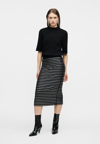 Γυναίκα που φοράει μαύρο μπλουζάκι με κοντά μανίκια, μαύρη μεσαίου μήκους φούστα με ασημένια κυκλικά σχέδια, μαύρα μποτάκια μέχρι τον αστράγαλο και κρεμαστά σκουλαρίκια, που στέκεται μπροστά σε λευκό φόντο.