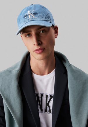 Denim cap with embroidered logo, light blue color; model wearing a black blazer, gray sweatshirt draped over shoulder, white t-shirt.