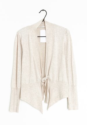 Cardigan beige clair à manches longues avec un nœud noué devant, poignets côtelés, épaules légèrement bouffantes, suspendu à un cintre noir sur fond blanc.