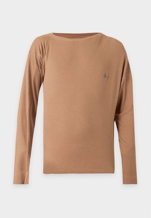 Man staat met handen in de zakken, draagt een bruine lange mouwen overhemd, geruite broek en bruine loafers tegen een effen achtergrond.