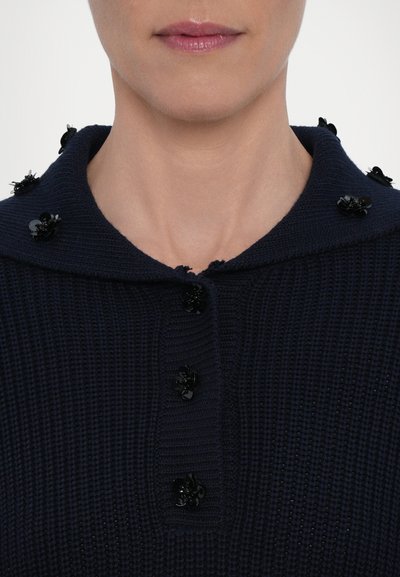 Marinblå stickad tröja med en stor krage, prydd med svarta blommiga dekorationer på kragen och knapparna. Texturerad tygfinish.