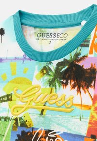 T-shirt a maniche corte in cotone organico azzurro chiaro con una vivace grafica di palme e sole, caratterizzata da un logo "Guess" ricamato in giallo.