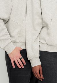 Deux personnes portant des sweat-shirts gris clair et des pantalons foncés se tiennent côte à côte, l'une ayant une bague argentée à la main gauche.