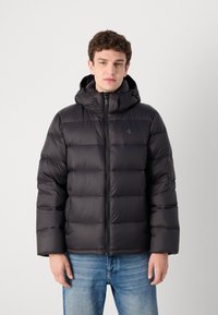 Black puffer jacket with a hood, featuring a zip front, quilted design, and logo detail on the chest. Worn over light blue jeans.