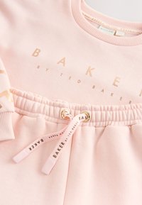 Gros plan sur un sweat-shirt rose pâle et un pantalon avec taille élastique, cordon avec texte "BAKER by Ted Baker" en lettres dorées.