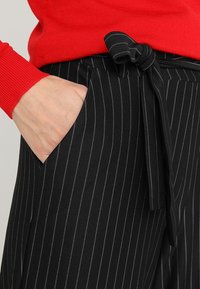 Pantalon rayé noir et blanc avec une ceinture nouée, doté de deux poches latérales. Le tissu semble lisse avec un léger brillant, assorti à un pull rouge.